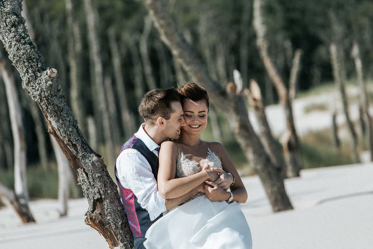 uśmiechnięta Pana młoda nad morzem oparta o drzewo 